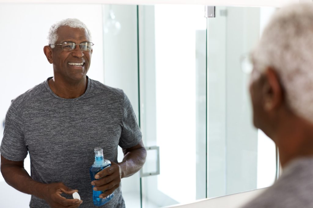 Senior Man Using Mouthwash Looking At Reflection In Bathroom Mirror Wearing Pajamas